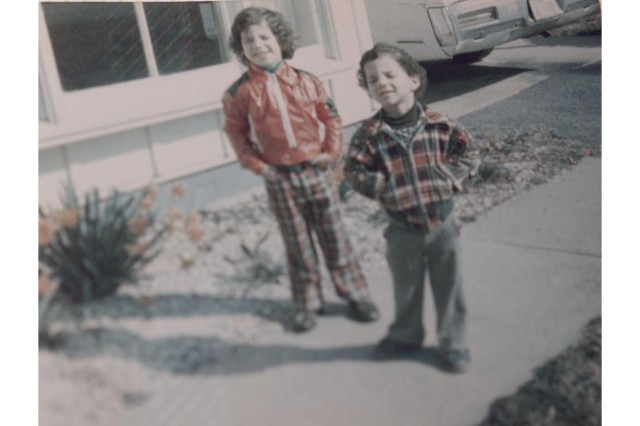 Two young boys in the late 1970s with short curly hair, squinting against the sun, standing on the sidewalk in front of their house, hands in their pockets, dressed in matching plaid jackets and pants. 